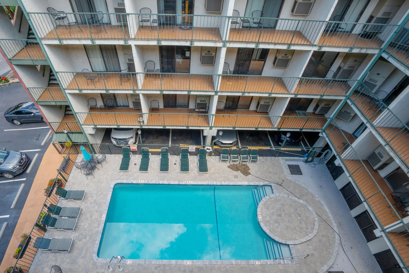 An aerial view of the property reveals a central swimming pool surrounded by lounge chairs, enclosed by multiple stories of accommodations with private balconies. Parking is conveniently located nearby.