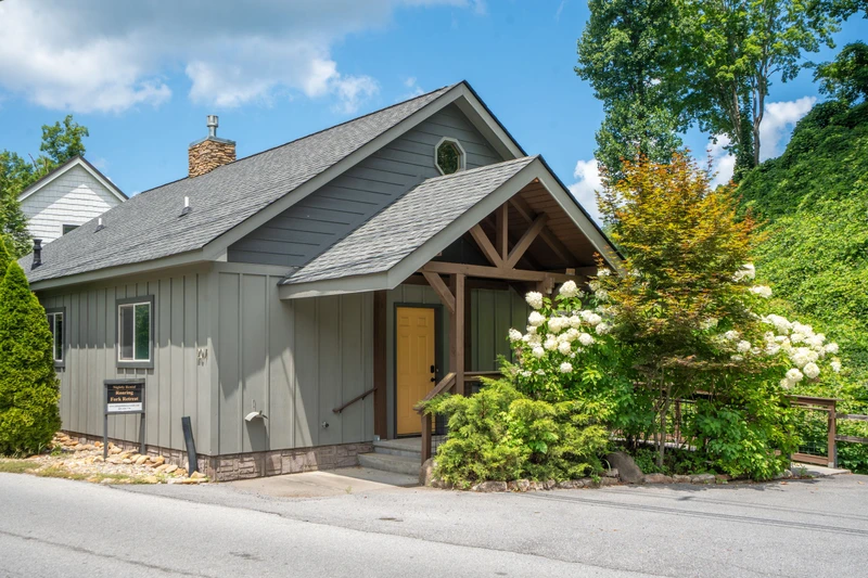 Charming gray cottage surrounded by lush landscaping and mature trees, featuring a welcoming covered entrance and abundant natural greenery in a peaceful setting.