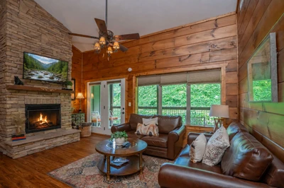 Settle into this warm, inviting living room with rich wood walls, a cozy fireplace, and leather seating perfect for gathering. Large glass doors open to your private deck overlooking lush greenery, creating your peaceful mountain retreat.