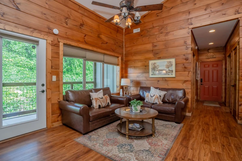 Relax in your cozy cabin living space with warm wood walls, leather sofas, and lush greenery framing the glass doors—your peaceful retreat after a day of adventure.