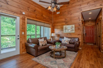 Relax in your cozy cabin living space with warm wood walls, leather sofas, and lush greenery framing the glass doors—your peaceful retreat after a day of adventure.