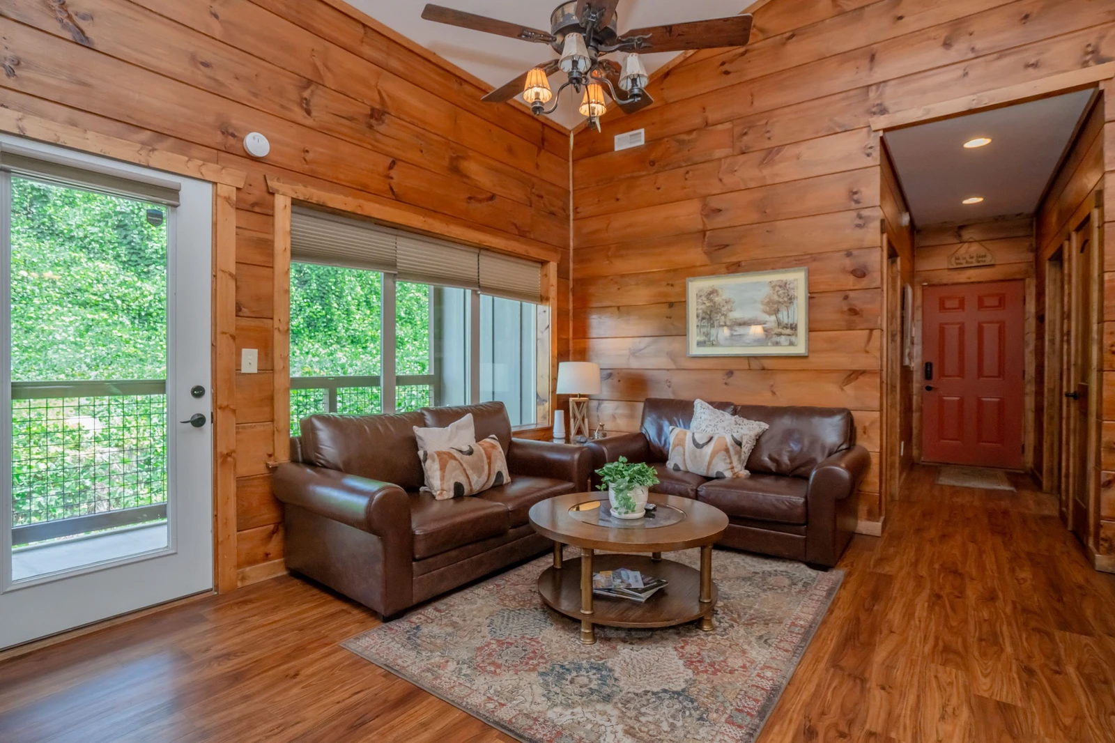 Relax in your cozy cabin living space with warm wood walls, leather sofas, and lush greenery framing the glass doors—your peaceful retreat after a day of adventure.