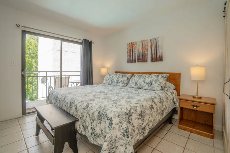 Relax in your cozy bedroom with soft bedding and natural light streaming through the balcony door. Step outside for a breath of fresh air or unwind with the peaceful wooded scene above the bed.
