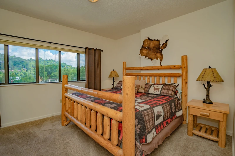 Your cozy mountain bedroom features handcrafted log bunk beds and charming rustic touches, while large windows frame stunning forested hillside views for a peaceful retreat.