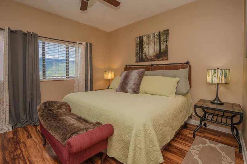 Unwind in your cozy bedroom featuring warm hardwood floors, a plush bed with soft linens, and natural light that frames peaceful mountain views through the window.