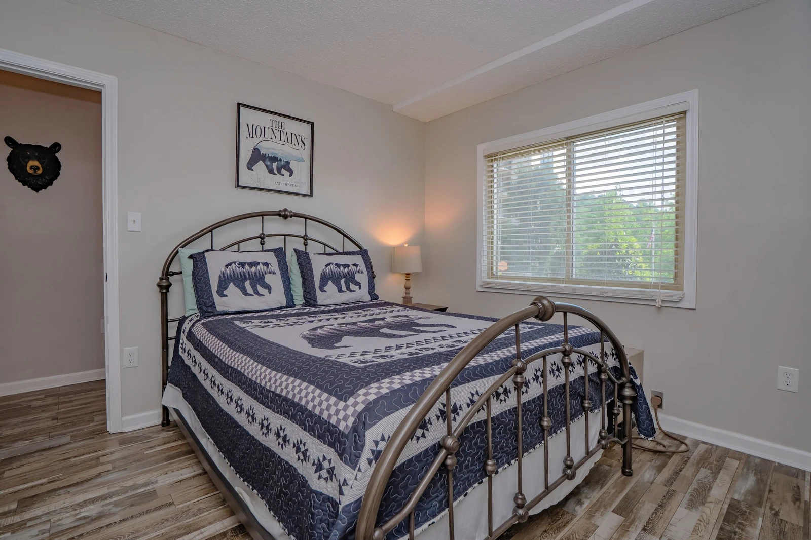 Relax in your cozy mountain-themed bedroom featuring a charming metal-frame bed dressed in rustic bear-motif linens, with natural light filtering through the blinds to brighten your morning.