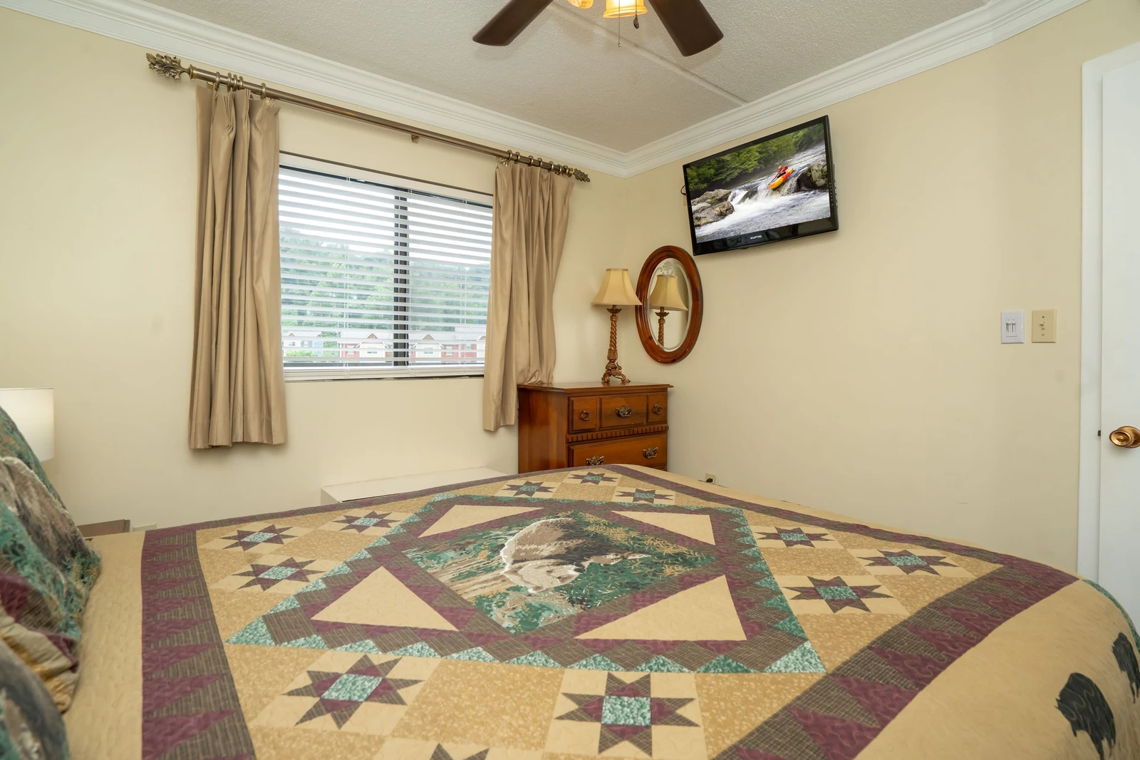 Your bedroom features a handsome quilt-topped bed and warm wooden furnishings, lit by soft natural light through the window and the gentle glow of a ceiling fan above.