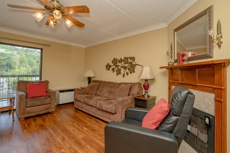 Settle in and unwind in this cozy living room with comfortable seating, warm wood accents, and a ceiling fan to keep you cool. Enjoy peaceful views through the window and the inviting glow of soft lighting.
