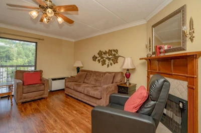 Settle in and unwind in this cozy living room with comfortable seating, warm wood accents, and a ceiling fan to keep you cool. Enjoy peaceful views through the window and the inviting glow of soft lighting.
