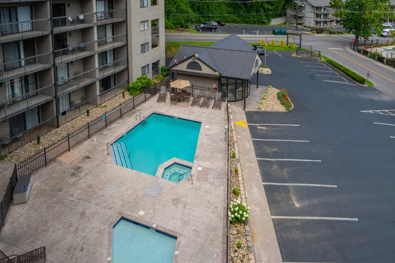 Aerial view of the property featuring an outdoor pool with covered gazebo seating area and ample guest parking adjacent to tree-lined streets and nearby buildings.