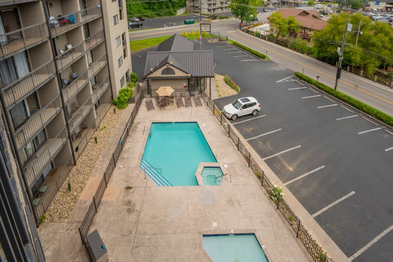 An aerial view of the property showcases the swimming pool, outdoor seating pavilion, and ample parking, with the surrounding residential neighborhood and green springtime landscape beyond.