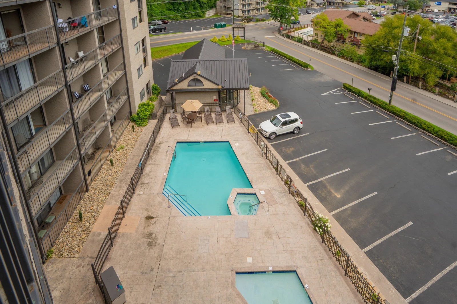 An aerial view of the property showcases the swimming pool, outdoor seating pavilion, and ample parking, with the surrounding residential neighborhood and green springtime landscape beyond.