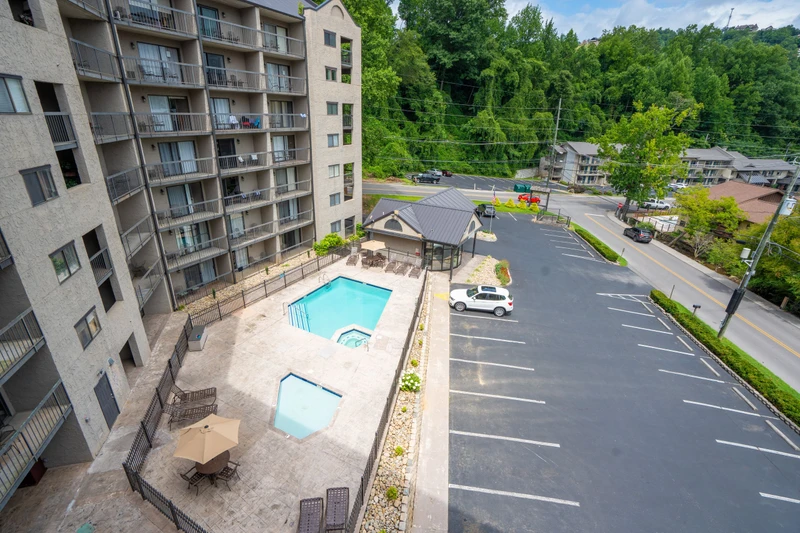 An aerial view shows the property nestled in a lush, wooded setting. The outdoor pool and parking area are conveniently located on-site, surrounded by dense summer foliage and nearby accommodations.