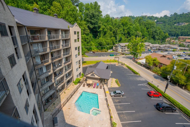 This aerial view captures the property nestled in a lush, tree-covered valley with mountain vistas. The pool and parking area are conveniently located, with nearby shops and restaurants just a short stroll away.