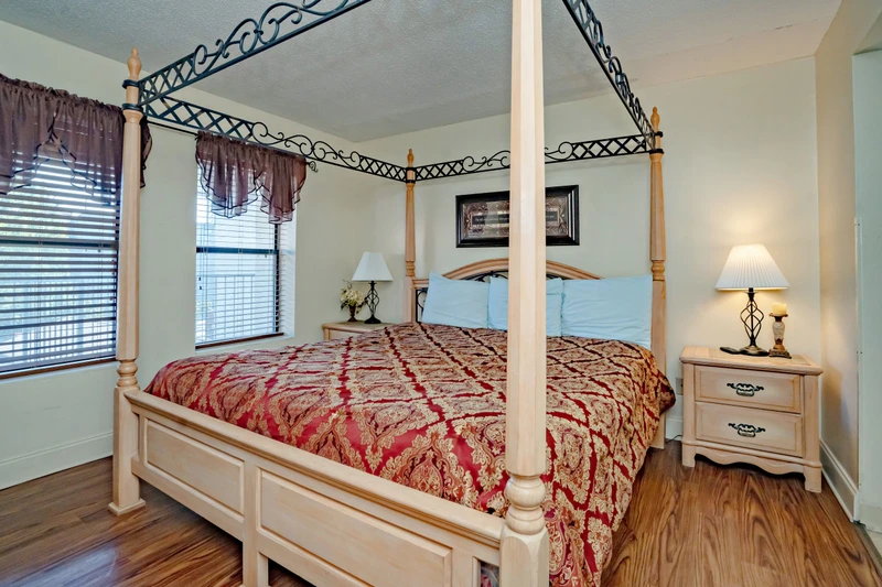 Step into your elegant bedroom featuring a romantic four-poster bed dressed in rich red and gold linens. Natural light filters through the windows while bedside lamps create a warm, inviting glow for your restful retreat.