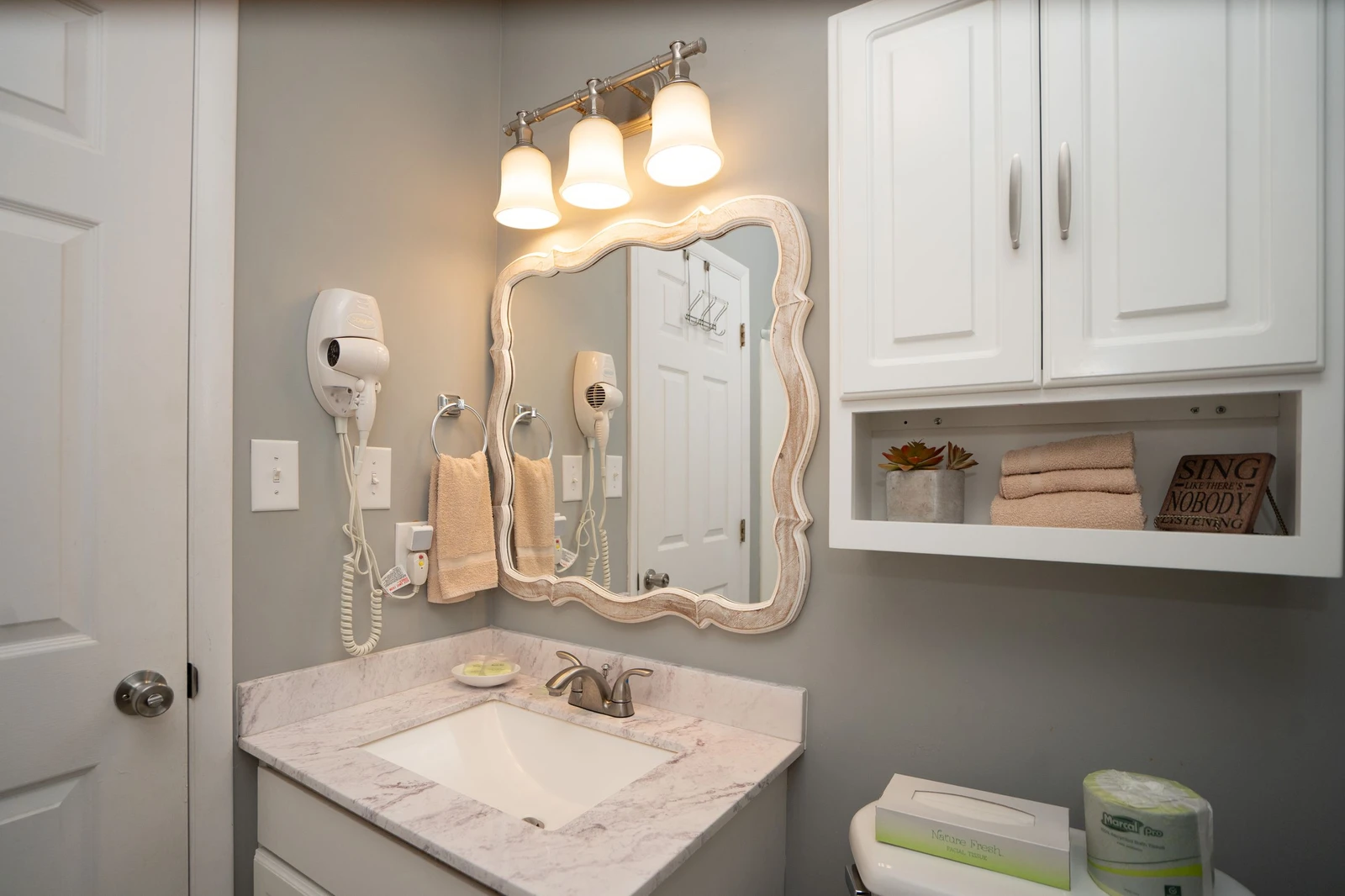 Freshen up in your charming bathroom, featuring soft lighting, a decorative mirror, and plush towels waiting for you. The marble vanity and thoughtful storage create a calming, well-appointed space for your comfort.